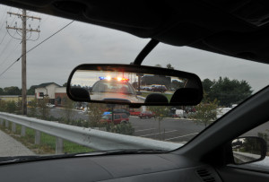 View from inside a car, looking through the rearview mirror, showing a police vehicle with flashing lights pulling over the car. The scene includes a street with parked cars and trees in the background, creating tension as thoughts of false Breathalyzer readings race through your mind.