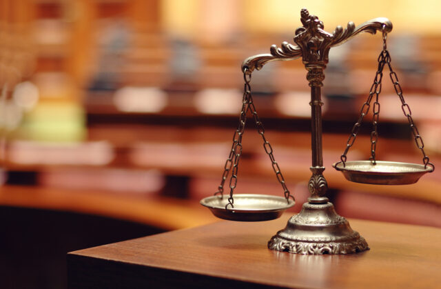 A bronze balance scale sits on a wooden desk in a courtroom. The scales are empty, and the background is blurred, with wooden seats and tables visible, suggesting a legal or judicial setting.