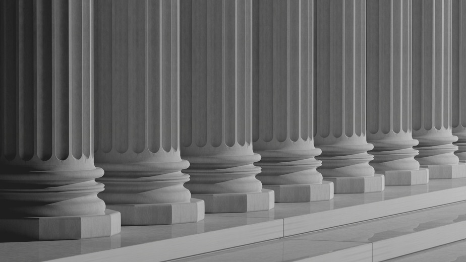 A row of classical Greek or Roman columns made of stone, shown from a low angle. The columns are fluted and evenly spaced, creating a sense of symmetry and elegance. The image is in grayscale, enhancing the architectural details and textures.