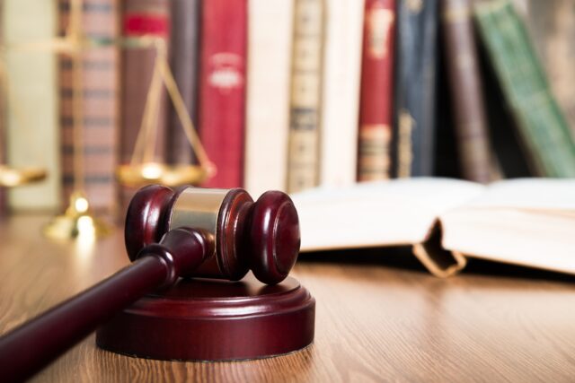 A judge's wooden gavel rests on a sound block on a desk. In the background, there are a balance scale and several legal books on a bookshelf, suggesting a legal or judicial setting.