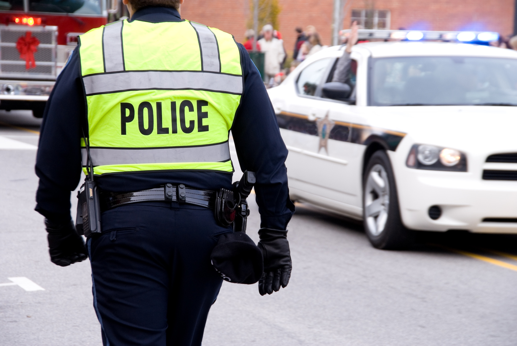 A police officer in a neon yellow safety vest stands on a street with a patrol car in the background. The officer appears to be managing traffic or a crowd, with people and a red brick building visible in the background.