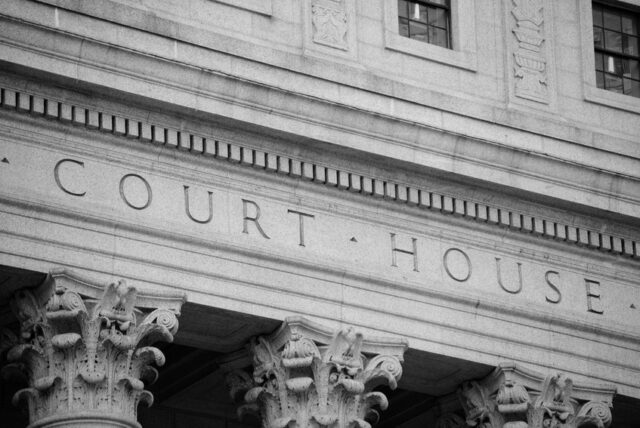 Black and white image of the exterior of a courthouse. The word "COURT HOUSE" is prominently inscribed on the building's facade above tall, ornate columns with intricate detailing. The architecture features classical design elements.