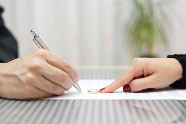 Two hands are depicted, one holding a pen and signing a document, and the other pointing at a ring placed on the document. The background is blurred, showing a green plant and curtains.