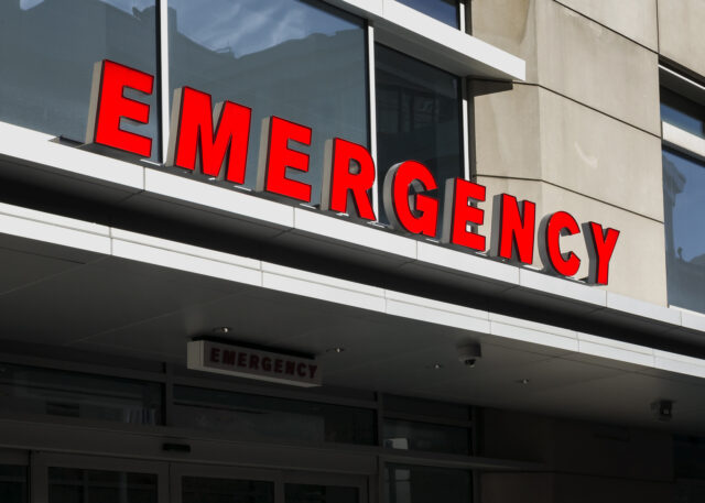 A building exterior with a large, bold, red "EMERGENCY" sign above the entrance, indicating the location of an emergency room at a hospital or medical facility. The architecture is modern with large windows and a stone facade.