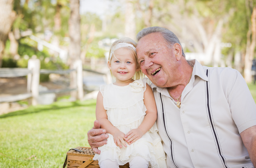 An elderly man and a young child, both smiling, sit close together outdoors on a sunny day. The man has gray hair and wears a light-colored shirt, while the child is dressed in a white outfit with a matching headband. A green, leafy background is visible behind them.