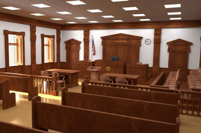 A courtroom with wooden benches, a judge's bench, and a witness stand. The room includes an American flag, a clock on the wall, and a seal in front of the judge's bench. There are large windows allowing natural light to enter the room.