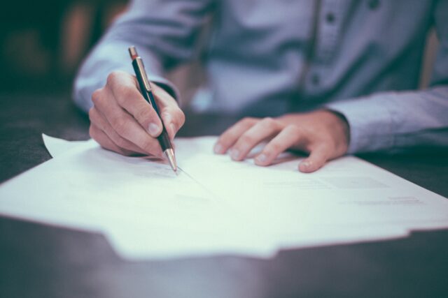 A person in a light blue shirt is seated and writing on several sheets of paper with a pen. Only their hands and part of their arms are visible in the image. The background is blurred, focusing attention on the writing activity.