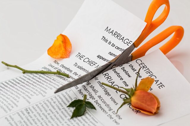 A pair of orange-handled scissors cutting through a marriage certificate. A yellow rose lies on the certificate, with one petal removed, which is also on the torn document.