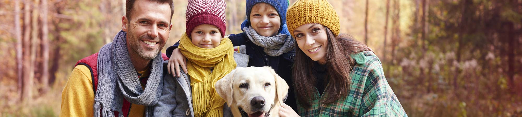 A family of four, with two adults and two children, poses outdoors in autumn attire, including colorful scarves and hats. They are smiling and standing close together with a golden retriever at the center. The background features trees with fall foliage.