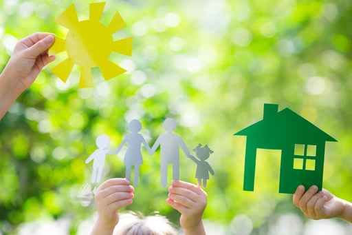 Hands holding paper cutouts of a sun, a family, and a house against a blurred green background. The cutouts depict a family of four, including two adults and two children, symbolizing concepts of home, family, and environmental consciousness.