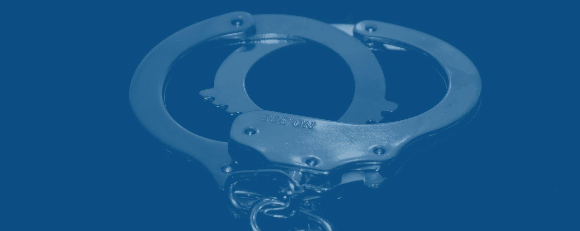 A pair of metal handcuffs is displayed against a dark background, with a blue filter overlay. The handcuffs are open and positioned in such a way that both cuffs are clearly visible.