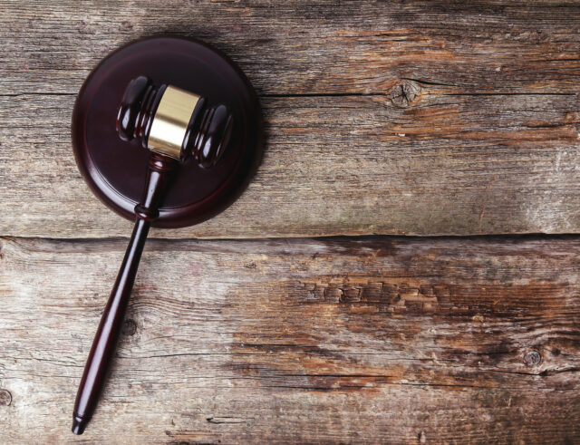 A wooden judge's gavel and sound block placed on a rustic wooden surface, symbolizing justice and authority in a judicial setting. The gavel is positioned on the left side of the image, with the handle extending towards the right.