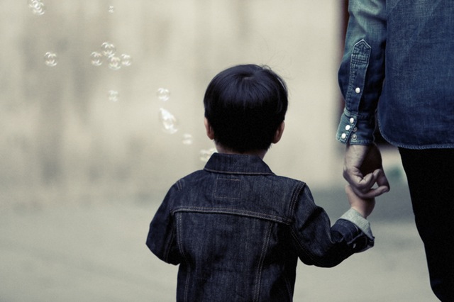 A child wearing a denim jacket holds hands with an adult, also in denim, while walking outdoors. The scene is accompanied by floating bubbles, creating a playful and serene atmosphere. The view is from behind, obscuring the identities of both individuals.