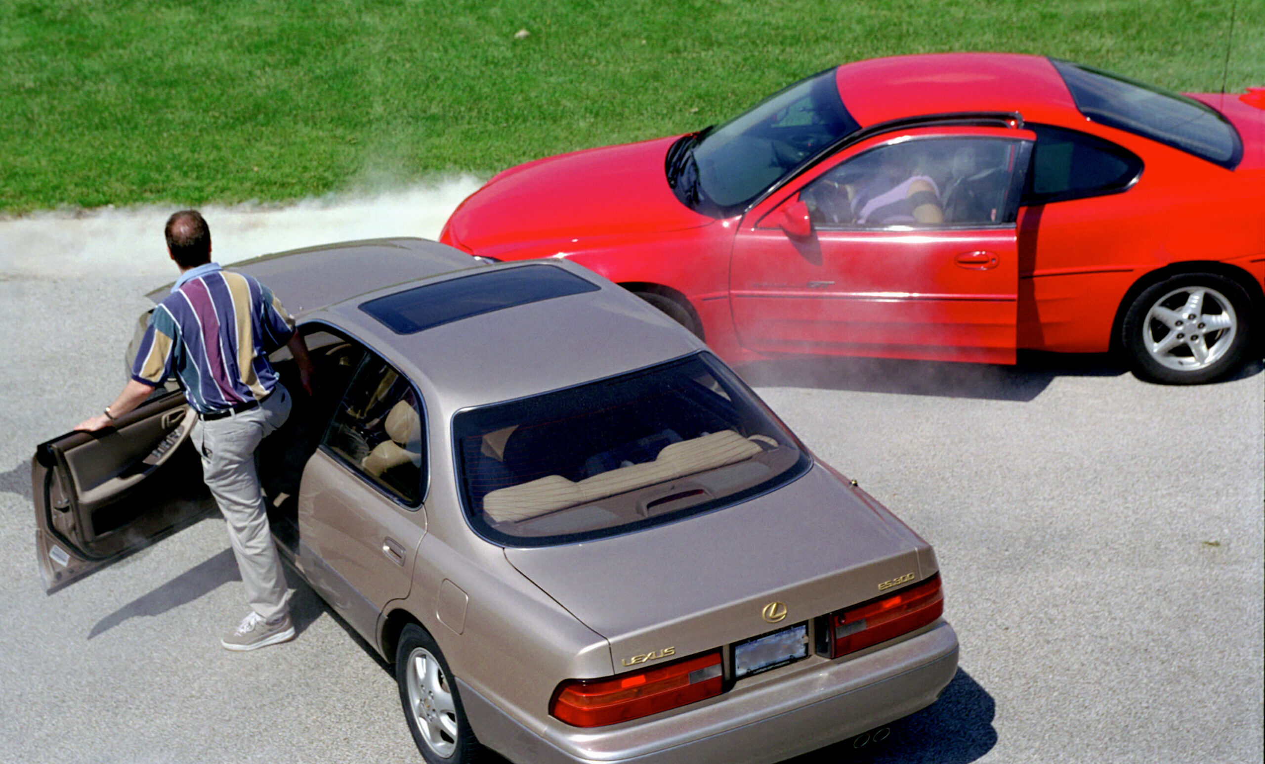 A man steps out of a beige Lexus car after a collision with a red car on a Pennsylvania road. The red car has smoke emanating from its hood and both cars show visible damage. Unsure about what to do next, the scene unfolds on a paved road with grass in the background.