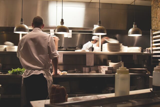 A busy restaurant kitchen in Pennsylvania with chefs in white uniforms. One chef with their back to the camera is preparing food at a counter, while others are seen plating dishes and handling utensils. Various plates, ingredients, and kitchen equipment are visible after a recent restaurant accident.