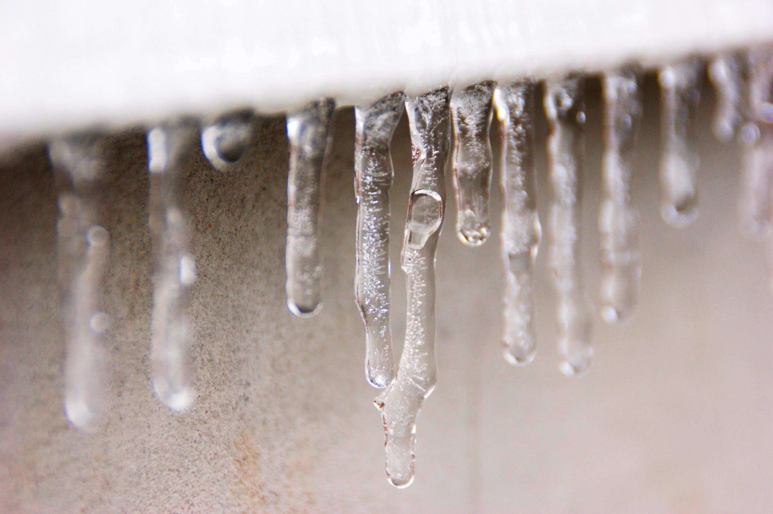 Close-up of icicles hanging from the edge of a surface, possibly a roof or eave. The icicles vary in length and shape, with some slightly blurred due to the depth of field. The background appears to be a concrete wall in Pennsylvania, where snow or ice accidents can lead to compensation claims.