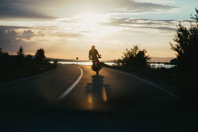 A person rides a motorcycle on a road toward a setting sun. The sky is filled with clouds, and the silhouette of the rider, who appears to be mindful of motorcycle helmet laws in Pennsylvania, is framed by trees and distant hills. The road is flanked by greenery, and the light casts a serene, warm glow over the scene.