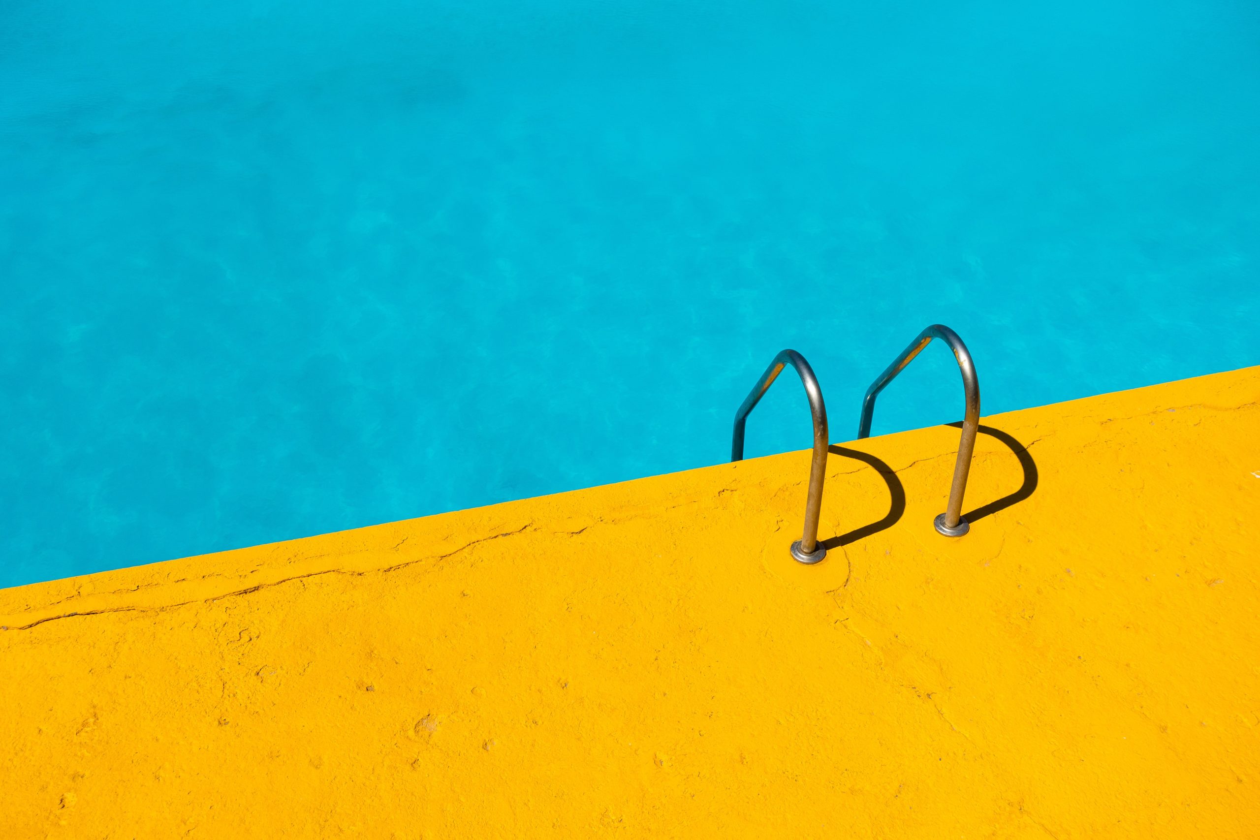 A vibrant swimming pool with crystal clear blue water and a bright yellow deck. A metal handrail for entering and exiting the pool is visible on the right side, contrasting with the intense yellow color of the deck. The reflection shimmers in the water, highlighting safety features to prevent pool accidents.