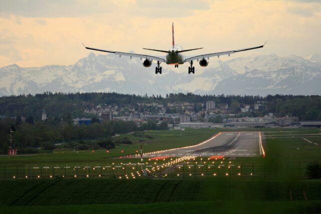 A commercial airplane is seen landing on a runway with illuminated lights at dusk, showcasing the safety measures in place to prevent airport accidents. In the background are snow-capped mountains and a city with various buildings surrounded by trees and greenery, beneath a partly cloudy sky.