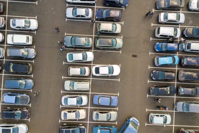 Aerial view of a crowded parking lot filled with various cars in organized rows. A few pedestrians cautiously walk between the parked vehicles. Some empty parking spaces can be seen among the occupied spots. The ground, paved with asphalt, shows no signs of any recent parking lot accident.