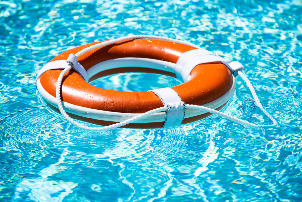 A bright orange life preserver ring with white straps and ropes floats on the sparkling blue water of a swimming pool. The sunlight reflects off the water, creating a vibrant and inviting scene, perfectly showcasing the allure of a summer day spent by the swimming pool.