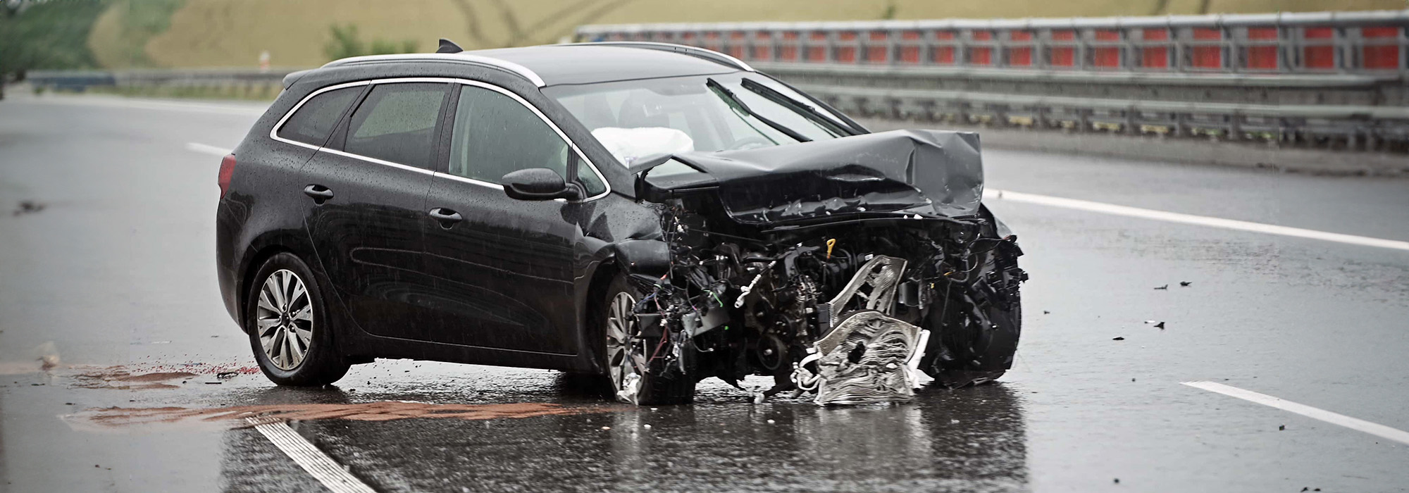 A black car with significant front-end damage is shown stopped on a wet highway. Airbags have deployed, and debris is scattered around. The road is empty with fields in the background and a crash barrier on the right side. Those involved may need to consult a Car Accident Attorney for assistance.