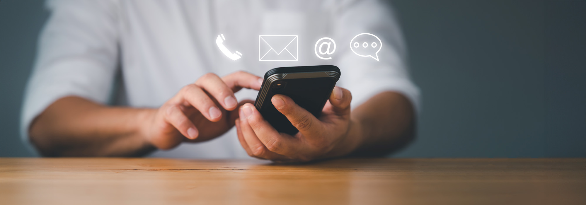 A person wearing a white shirt is using a smartphone at a wooden table. Above the phone, holographic icons of a phone call, email, internet (