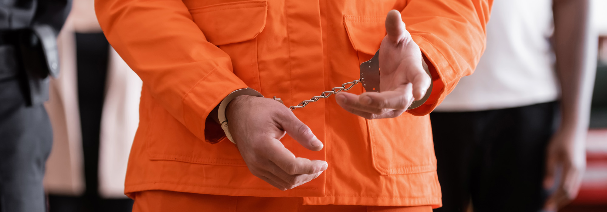 A person wearing an orange jumpsuit is shown from the shoulders down with their hands cuffed in front of them, connected by a chain. Other blurred figures can be seen in the background, possibly including an expungement lawyer focused on resolving their case.