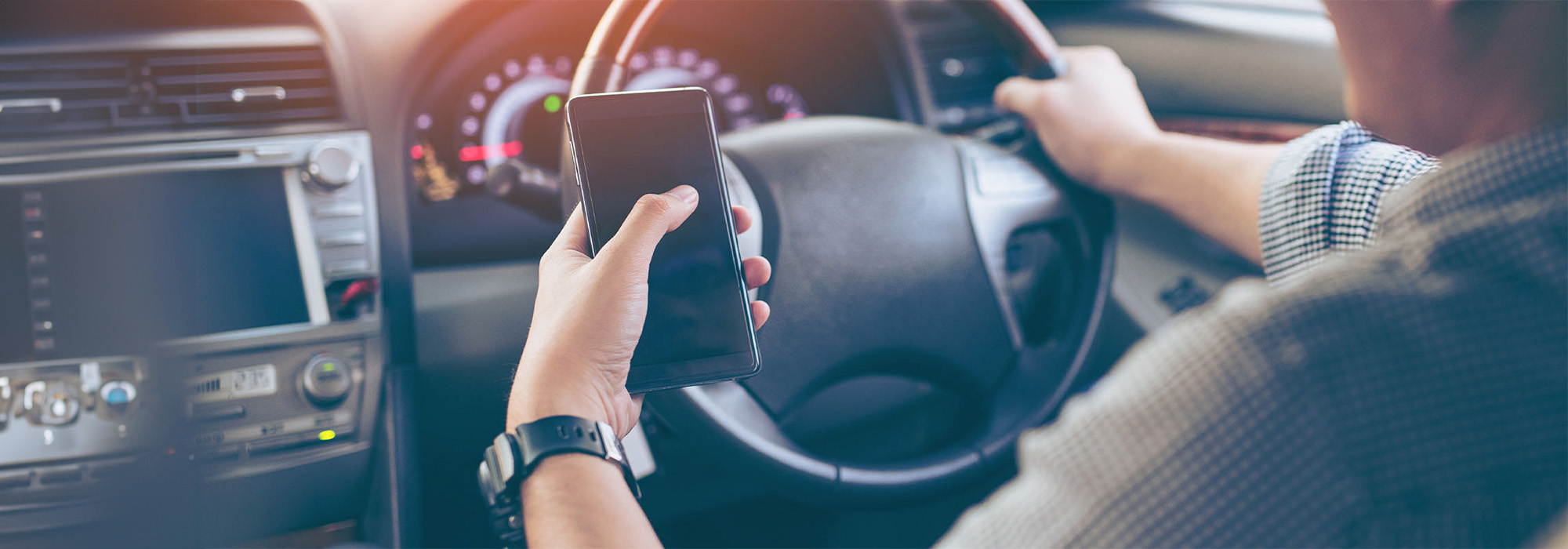 A person is driving a car while holding a smartphone with their right hand, clearly a distracted driver. The dashboard, steering wheel, and part of the infotainment system are visible. The person's left hand is on the steering wheel, and they are wearing a wristwatch on their left wrist.