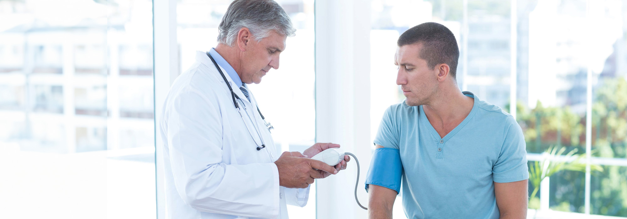 A physician, donned in a white coat, checks a patient's blood pressure with a monitor in a bright medical office. The patient, seated and wearing a blue armband, looks concerned while the doctor examines the screen of the device. This scene reflects physician representation at its finest.