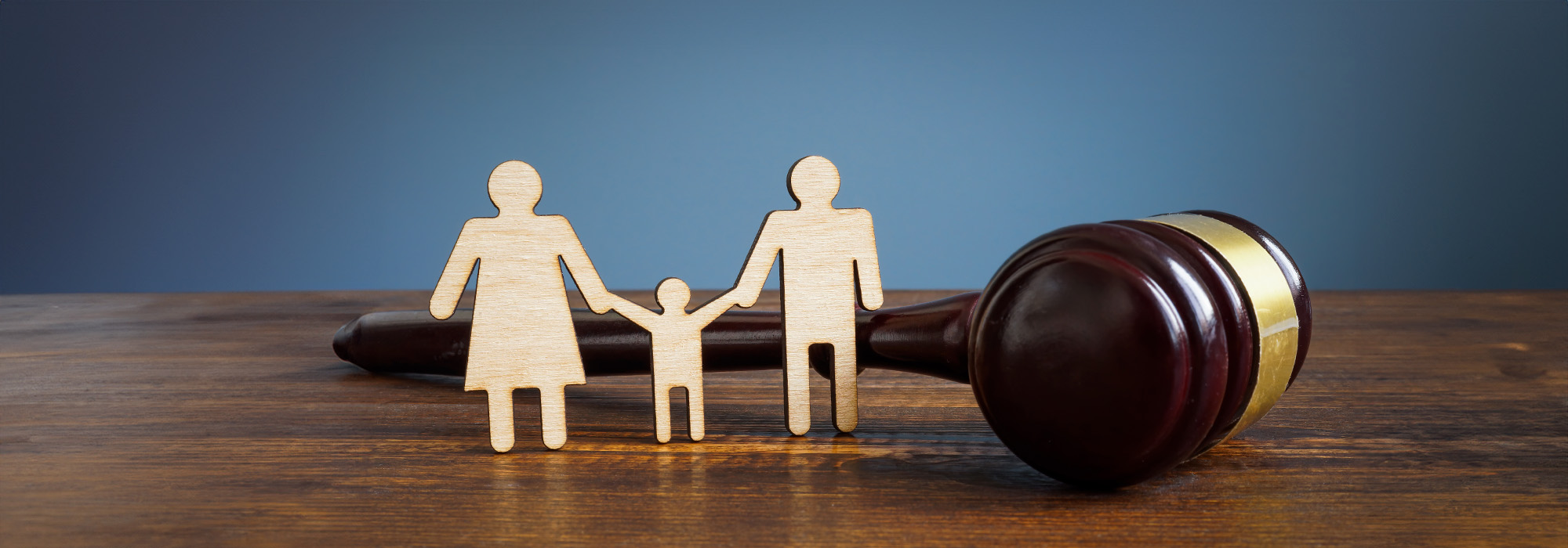 A wooden cutout of a family consisting of two adults and one child stands in front of a judge's gavel on a wooden surface. The background is a gradient of blue. This image symbolizes family law or legal matters related to families, an area diligently handled by York County Family Law Attorneys.