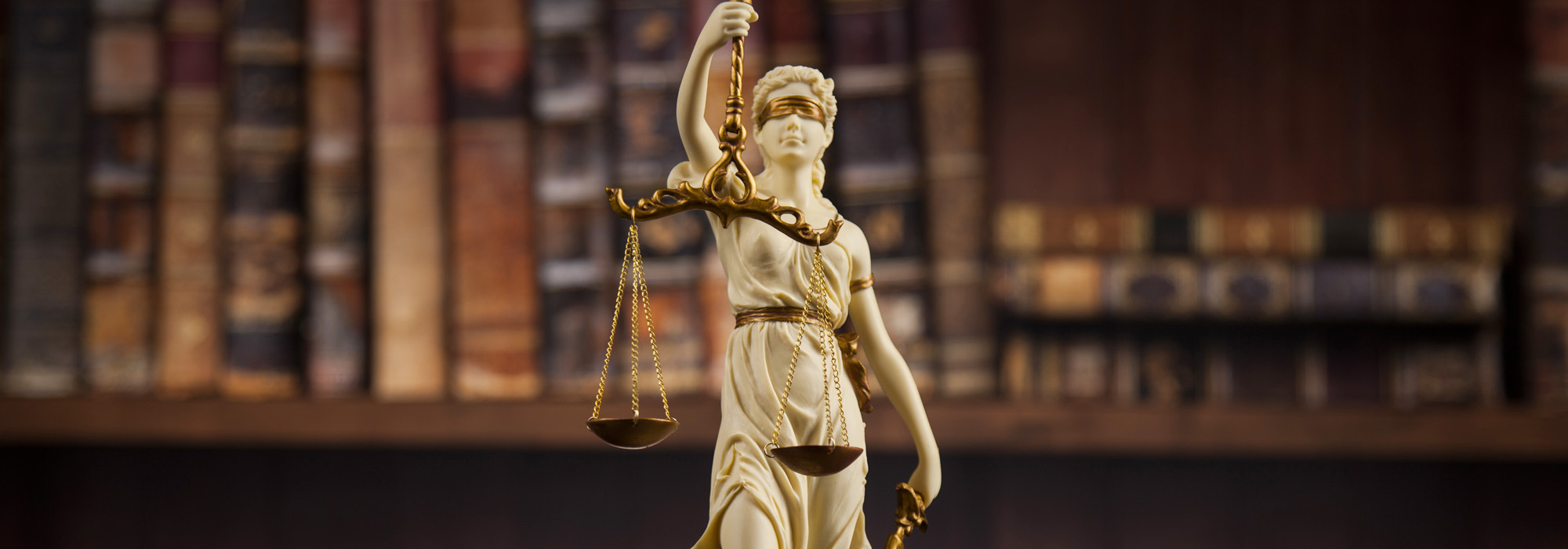 A statue of Lady Justice holding scales stands prominently in front of a blurred background of bookshelves. The statue, symbolizing the core tenets of legal services, features a blindfold representing impartiality and holds scales to denote fairness and balance in the justice system.