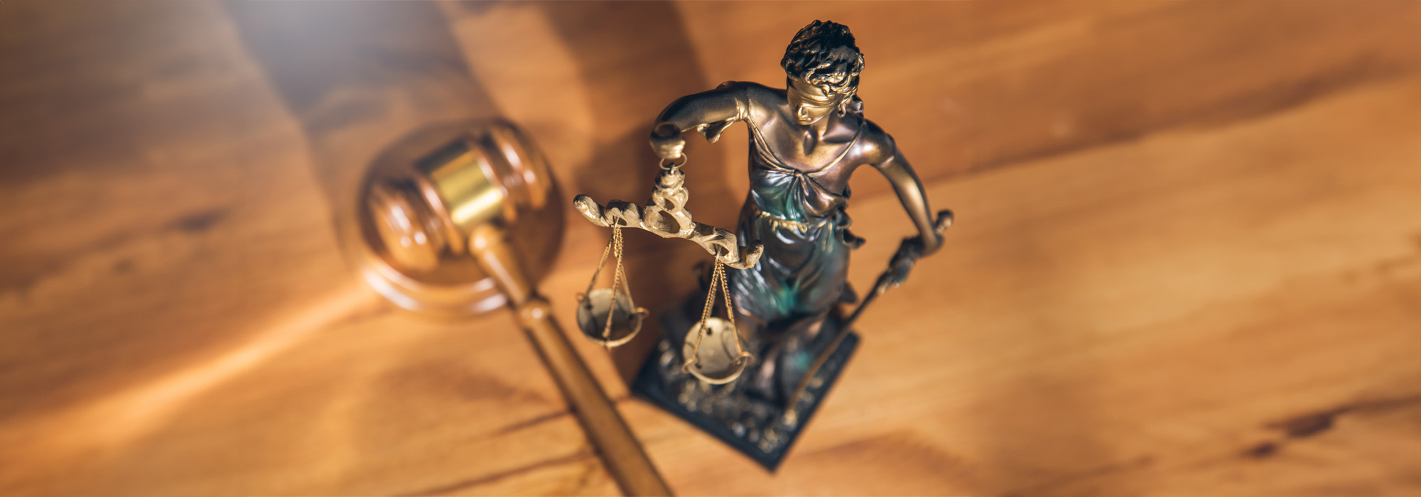 A bronze statue of Lady Justice is shown, featuring scales in one hand and a sword in the other. The statue is placed on a wooden surface next to a gavel, symbolizing various practice areas of law and justice. The image has a soft light highlighting the objects.