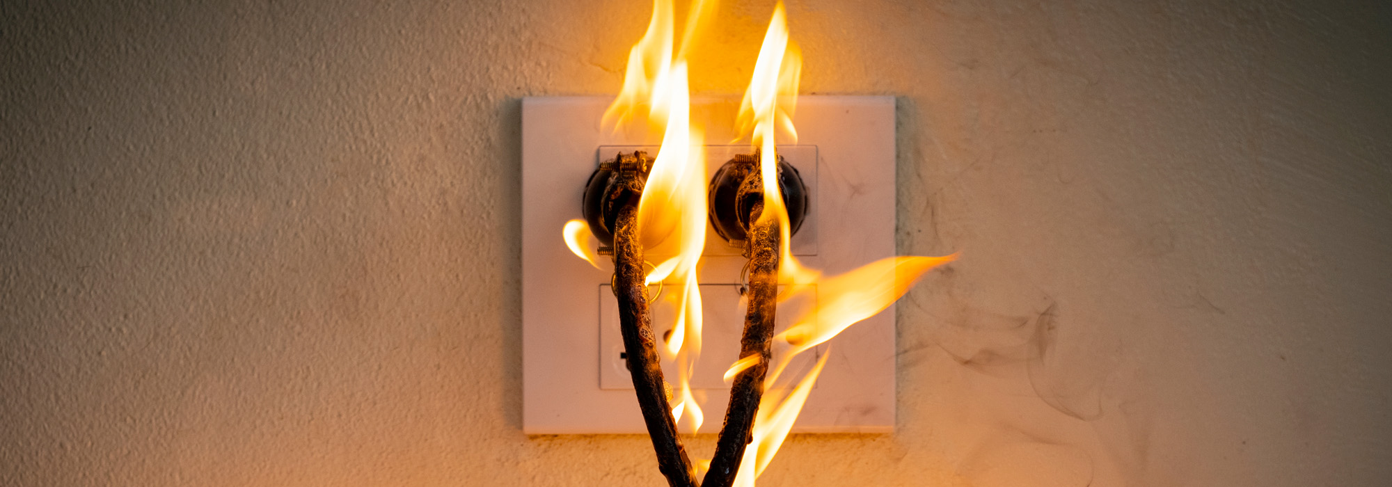 A close-up image shows an electrical outlet on fire. Two plugged-in cables blaze with flames and smoke against the wall, signaling a hazardous situation. The background wall is slightly scorched from the heat, highlighting the severity of potential dangers that might require a burn injury attorney.