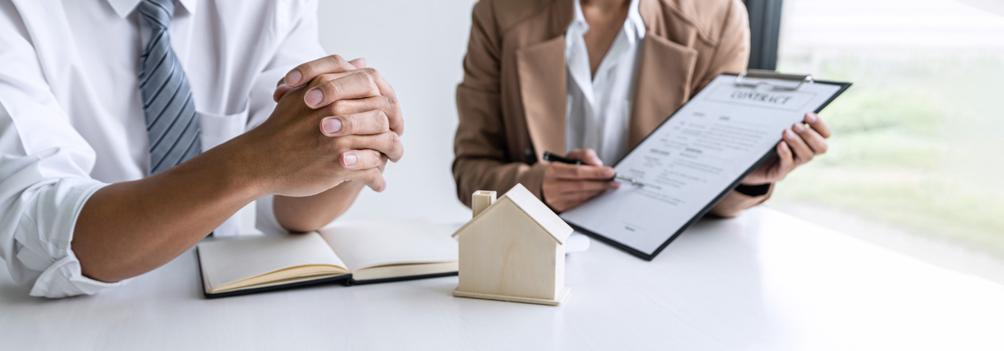 A person in a suit holds a clipboard with a visible contract, while another individual with clasped hands sits across the table. A small wooden house model and an open notebook on the table suggest a meeting involving estate planning attorneys or real estate business.