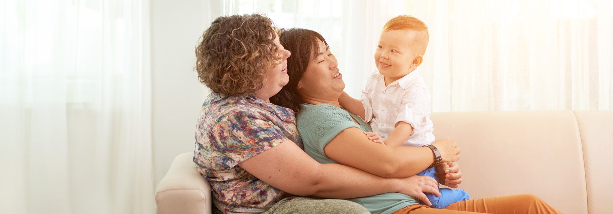 A family sitting on a couch in a cozy, sunlit room. Two adults smile while holding a happy young child in a white shirt and blue pants. One adult with curly hair wears a patterned shirt, the other adult with straight hair wears a green top, showcasing the joy that LGBTQ Family Law Attorneys strive to protect.